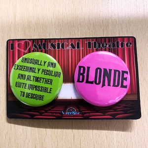 Puede incluir: Dos botones redondos con fondo rojo, blanco y negro. El botón verde dice "Unusually and Exceedingly Peculiar and Altogether Quite Impossible to Describe". El botón rosa dice "Blonde".