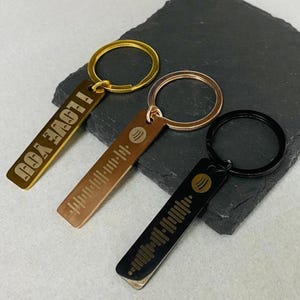May include: Three metal keyrings in gold, bronze and black. One gold keyring has the text "I LOVE YOU". The other two keyrings have a Spotify code design.