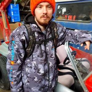 May include: A person wearing a camouflage jacket with an orange beanie and a black backpack. The jacket has the text "ACTIVE" printed on the sleeve. The person is standing next to a red and silver vehicle.