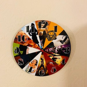 May include: A round clock with a colorful design featuring images from popular musicals. The clock face is divided into 12 sections, each with a different musical theme. The numbers 1 through 12 are displayed in black on a white background. The clock is made of a durable material and is perfect for any Broadway fan.