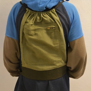 May include: A person wearing a blue hoodie with brown sleeves and a green backpack. The backpack has a large pocket with a zipper. The person is also wearing a navy blue skirt.