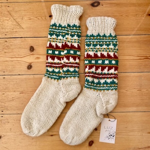 May include: A pair of cream-coloured knitted socks with a patterned design in green, burgundy, and gold. The socks have a ribbed cuff and a decorative band of geometric shapes. A tag with the brand name is attached.