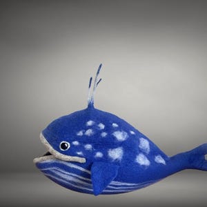May include: A handcrafted blue whale plush toy with white spots and a fin-like tail. The whale has an open mouth and a small fin on top of its head. The toy is made of soft material and is suitable for children.