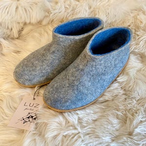 May include: A pair of grey felt slippers with a blue interior and a light brown sole. The slippers are displayed on a fluffy, white surface. A tag with the text "LUZ" is attached to one slipper.