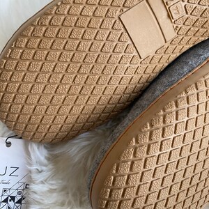 Puede incluir: Primer plano de la parte inferior de dos zapatillas de fieltro gris con suelas de goma marr&oacute;n. Las suelas tienen un patr&oacute;n de diamante. Las zapatillas est&aacute;n sobre una superficie blanca y esponjosa.