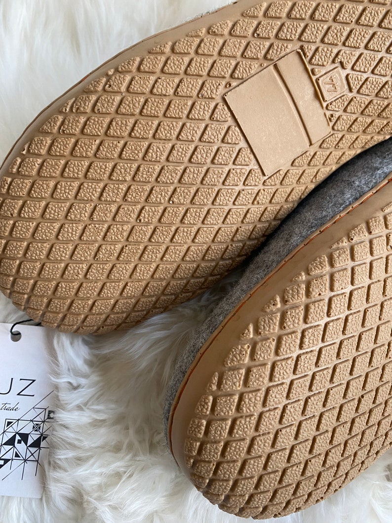 Puede incluir: Primer plano de la parte inferior de dos zapatillas de fieltro gris con suelas de goma marr&oacute;n. Las suelas tienen un patr&oacute;n de diamante. Las zapatillas est&aacute;n sobre una superficie blanca y esponjosa.