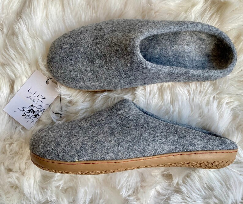 Puede incluir: Un par de zapatillas de fieltro gris con suela de cuero marr&oacute;n. Las zapatillas est&aacute;n sobre una superficie blanca y esponjosa. El nombre de la marca "LUZ" es visible en una etiqueta.
