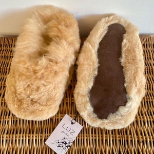Op de afbeelding: Een paar beige schapenvacht slippers met bruine su&egrave;de zolen. De slippers liggen op een geweven rieten oppervlak. Het label op de slippers luidt "Luz, your style".