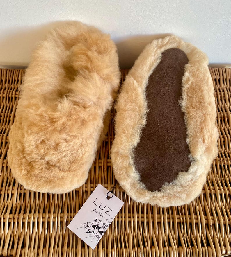 Op de afbeelding: Een paar beige schapenvacht slippers met bruine su&egrave;de zolen. De slippers liggen op een geweven rieten oppervlak. Het label op de slippers luidt "Luz, your style".