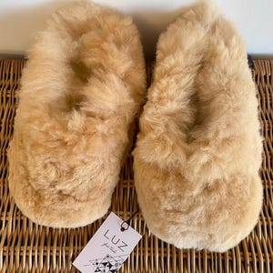 Op de afbeelding: Een paar beige wollen pantoffels met een pluche textuur. De pantoffels liggen op een geweven rieten oppervlak. Een label met de tekst "LUZ fair trade" is zichtbaar.