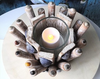 Puede incluir: Una vela blanca se encuentra dentro de un portavelas de vidrio rodeado por una disposición circular de madera flotante y conchas marinas. La madera flotante está pintada de blanco y las conchas marinas son azules y grises.