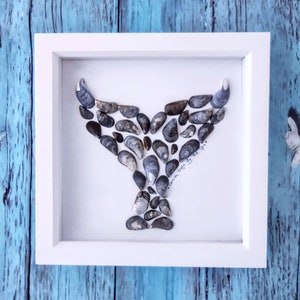 May include: A white framed picture with a whale tail made of seashells. The text "Taken Off By The Sea" is written in black ink on the white background.