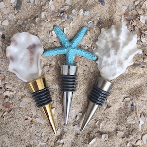 May include: Three decorative bottle stoppers with a beach theme. One stopper features a blue starfish, another a white seashell, and the third a white oyster shell. Each stopper has a silver metal base and a black rubber stopper.