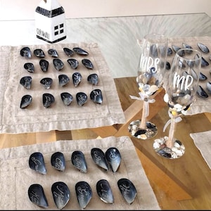 May include: A set of black and white seashell place cards with names written on them. Two champagne flutes with the words "Mrs." and "Mr." written on them are filled with small white and brown stones.