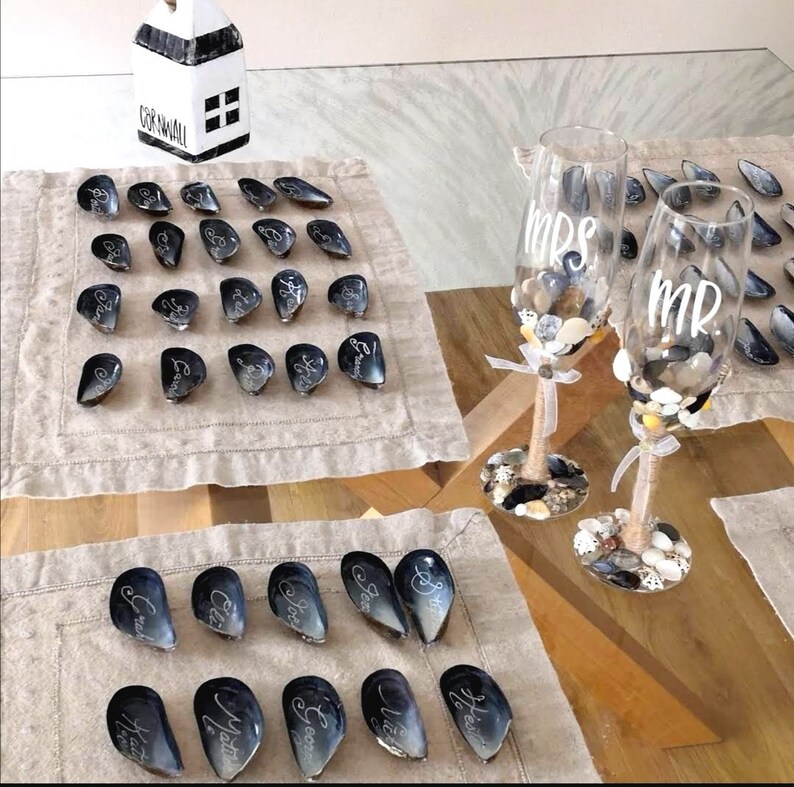 May include: A set of black and white seashell place cards with names written on them. Two champagne flutes with the words "Mrs." and "Mr." written on them are filled with small white and brown stones.