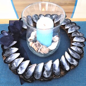 May include: A blue candle in a glass jar surrounded by a wreath made of mussel shells. The wreath is decorated with a black ribbon.