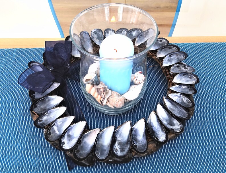 May include: A blue candle in a glass jar surrounded by a wreath made of mussel shells. The wreath is decorated with a black ribbon.