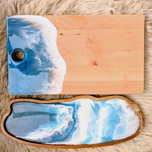 May include: Two wooden serving boards. One is rectangular with a blue and white resin wave design on one side and a hole for hanging. The other is an oval tray with a similar wave design. Both have a natural wood finish.