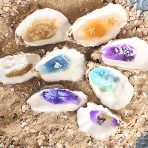 May include: Eight oyster shells filled with different coloured resin. The resin is clear, blue, purple, and gold. The shells are arranged on a sandy surface.