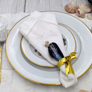 May include: A white napkin with a gold ribbon tied around it. The napkin is placed on a white plate with a gold rim. A black slate place card with the name "Becky" written on it is tucked into the napkin. The place setting is on a white tablecloth with a seashell design.