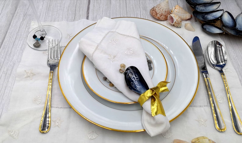 May include: A white napkin with a gold ribbon tied around it. The napkin is placed on a white plate with a gold rim. A black slate place card with the name "Becky" written on it is tucked into the napkin. The place setting is on a white tablecloth with a seashell design.
