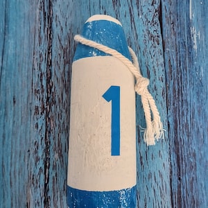 May include: A cylindrical wooden lobster buoy painted with a white centre and blue top and bottom sections. The number 1 is printed in blue on the white centre. A thick, off-white twisted rope is knotted around the top of the buoy.