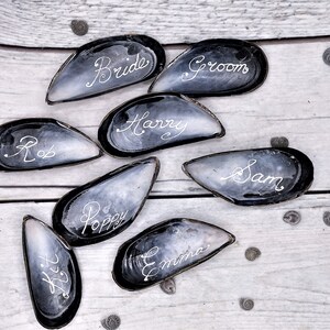 May include: Eight black mussel shells with white calligraphy on a white wooden surface. The names on the shells are Bride, Groom, Harry, Sam, Rob, Poppy, Emma, and  Kit.