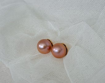 May include: A pair of round, blush-colored pearl stud earrings with gold-colored settings. The earrings are displayed on a white, sheer fabric background, creating a soft and elegant aesthetic. The pearls have a subtle luster.