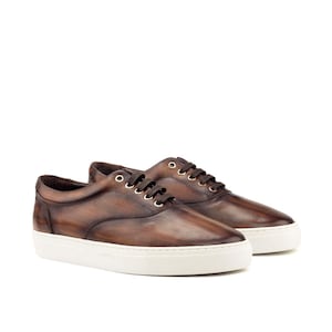 May include: A pair of low-top sneakers featuring a burnished brown leather upper with a distinct dark-to-light gradient finish. The shoes include dark brown laces, metallic eyelets, and a thick, solid white rubber sole with a textured sidewall.
