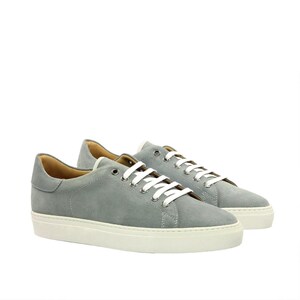 May include: A pair of low-top sneakers crafted from light gray suede leather. These shoes feature white flat laces threaded through silver-toned metal eyelets, a tan interior lining, and a thick, solid white rubber sole with a textured base.