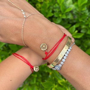 Puede incluir: Una colección de pulseras que incluye una cadena dorada con piedras claras, dos pulseras de hilo rojo con dijes de ojo dorados, un brazalete dorado con motivos de tornillos y una pulsera plateada con piedras claras y un ojo azul.