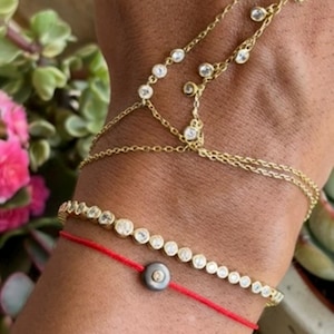 Op de afbeelding: Een gouden handketting met kleine in zettingen geplaatste heldere stenen, een gouden tennisarmband met een doorlopende rij heldere stenen, en een rood koordarmbandje met een donkergrijze ronde kraal met een kleine ingebedde steen.