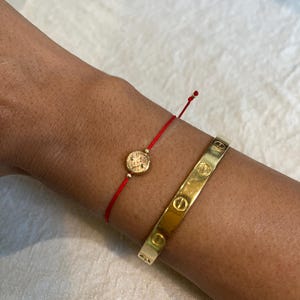 Puede incluir: Una pulsera de cordón rojo fino con un pequeño dije de moneda redondo en tono dorado con un perfil en relieve, junto a un brazalete rígido de metal pulido en tono dorado que muestra una serie de motivos de cabeza de tornillo espaciados.