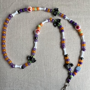 Puede incluir: Un cordón de cuentas con un patrón repetitivo de cuentas redondas moradas, naranjas y verde lima, intercaladas con cuentas blancas en forma de hueso, dijes negros con forma de murciélago y cuentas redondas diseñadas como globos oculares.