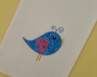 May include: A white kitchen towel with a blue bird embroidered on it. The bird has a pink heart on its wing and yellow feet.