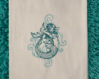 May include: A white kitchen towel with an embroidered teal mermaid design. The mermaid is curled up with her tail in a wave pattern.