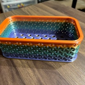 May include: A small, rectangular basket with a woven pattern. The basket is made of plastic and has a rainbow color scheme, with orange, green, and purple stripes.