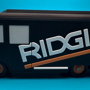 K&ouml;nnte beinhalten: Schwarzer Spielzeug-LKW mit dem Wort "RIDGID" in Wei&szlig; und Orange. Der LKW hat ein braunes Oberteil und orangefarbene Akzente an den R&auml;dern und der Sto&szlig;stange. Der LKW steht auf einer blauen Oberfl&auml;che.