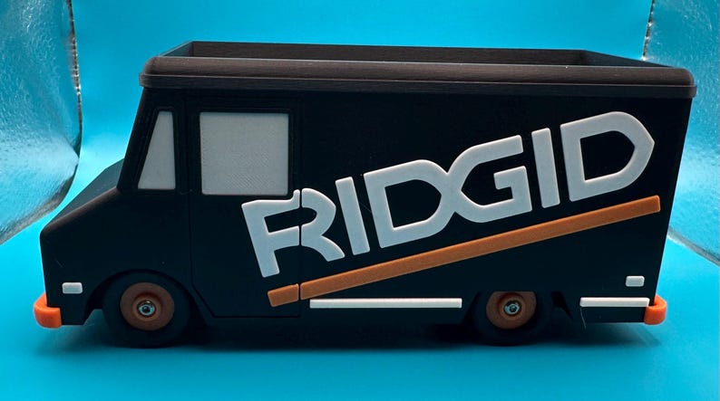 K&ouml;nnte beinhalten: Schwarzer Spielzeug-LKW mit dem Wort "RIDGID" in Wei&szlig; und Orange. Der LKW hat ein braunes Oberteil und orangefarbene Akzente an den R&auml;dern und der Sto&szlig;stange. Der LKW steht auf einer blauen Oberfl&auml;che.