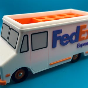 May include: A white FedEx Express truck-shaped desk organizer with orange and blue accents. The truck has black wheels, windows, and the FedEx logo on the side. The top of the truck is orange and has several compartments.