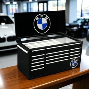 May include: Black toolbox-style cooler with the BMW logo on the lid and side. The cooler has white accents and a compartment with white inserts. It sits on a wooden table in a showroom.