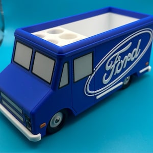 May include: A blue Ford truck-shaped desk organizer with a white interior and multiple compartments. The Ford logo is prominently displayed on the side. The organizer has a white front bumper and black wheels.