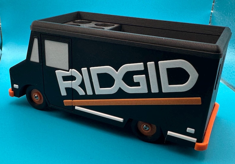K&ouml;nnte beinhalten: Ein schwarz-oranger Ridgid-Organizer in Form eines Lastwagens. Der LKW hat wei&szlig;e Schriftz&uuml;ge und orangefarbene Akzente. Die R&auml;der und Sto&szlig;stangen sind orange. Die Oberseite des LKWs hat mehrere kreisf&ouml;rmige F&auml;cher.