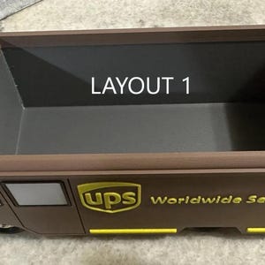K&ouml;nnte beinhalten: Ein brauner Container in Form eines UPS-Trucks mit dem UPS-Logo und "Worldwide Service" in goldfarbenen Buchstaben. Der Text "LAYOUT 1" ist im Inneren des Containers sichtbar. Er hat eine rechteckige &Ouml;ffnung.