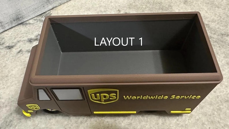 K&ouml;nnte beinhalten: Ein brauner Container in Form eines UPS-Trucks mit dem UPS-Logo und "Worldwide Service" in goldfarbenen Buchstaben. Der Text "LAYOUT 1" ist im Inneren des Containers sichtbar. Er hat eine rechteckige &Ouml;ffnung.
