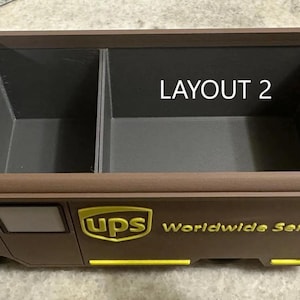 K&ouml;nnte beinhalten: Ein brauner und gelber UPS-Truck-f&ouml;rmiger Organizer mit zwei F&auml;chern. Der Text "LAYOUT 2" ist im Inneren des Organizers sichtbar. Das UPS-Logo und "Worldwide Service" sind auf der Seite aufgedruckt.