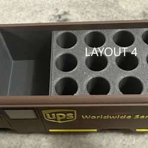 K&ouml;nnte beinhalten: Ein brauner UPS-LKW-f&ouml;rmiger Beh&auml;lter mit einem Staufach und einem Abschnitt mit kreisf&ouml;rmigen L&ouml;chern. Der Text "LAYOUT 4" ist sichtbar. Das UPS-Logo und "Worldwide Service" sind gelb gedruckt.