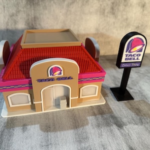 May include: A miniature Taco Bell restaurant model with a red roof, tan walls, and pink trim. The sign reads "TACO BELL" and includes a drive-thru sign. The model is on a white base.