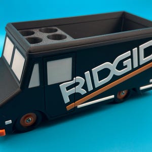 K&ouml;nnte beinhalten: Schwarzer und orangefarbener Schreibtisch-Organizer in Form eines Ridgid-Lastwagens. Der Organizer hat einen rechteckigen Stauraum und vier kreisf&ouml;rmige L&ouml;cher. Das Wort "RIDGID" ist in Wei&szlig; mit orangefarbenen Akzenten auf der Seite des Lastwagens dargestellt.