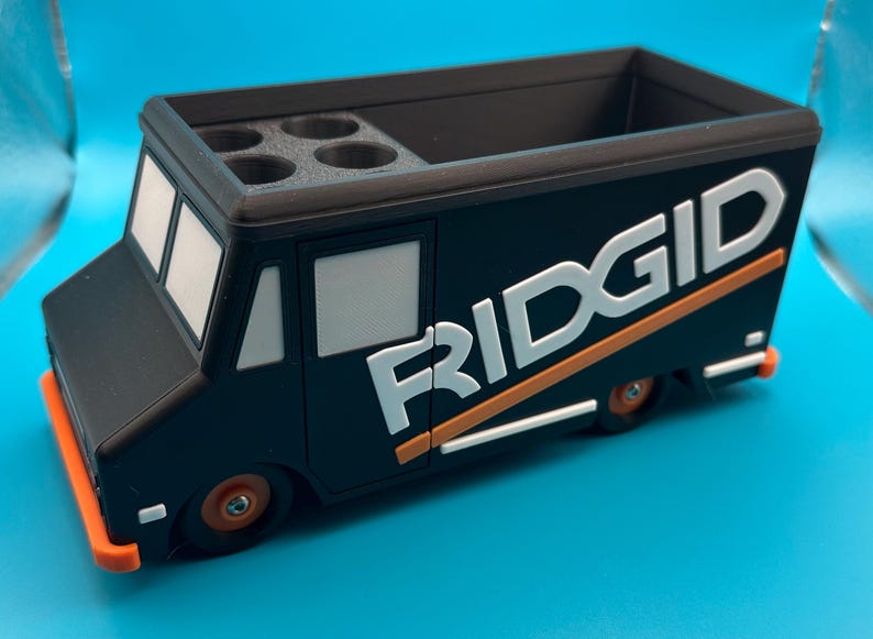 K&ouml;nnte beinhalten: Schwarzer und orangefarbener Schreibtisch-Organizer in Form eines Ridgid-Lastwagens. Der Organizer hat einen rechteckigen Stauraum und vier kreisf&ouml;rmige L&ouml;cher. Das Wort "RIDGID" ist in Wei&szlig; mit orangefarbenen Akzenten auf der Seite des Lastwagens dargestellt.